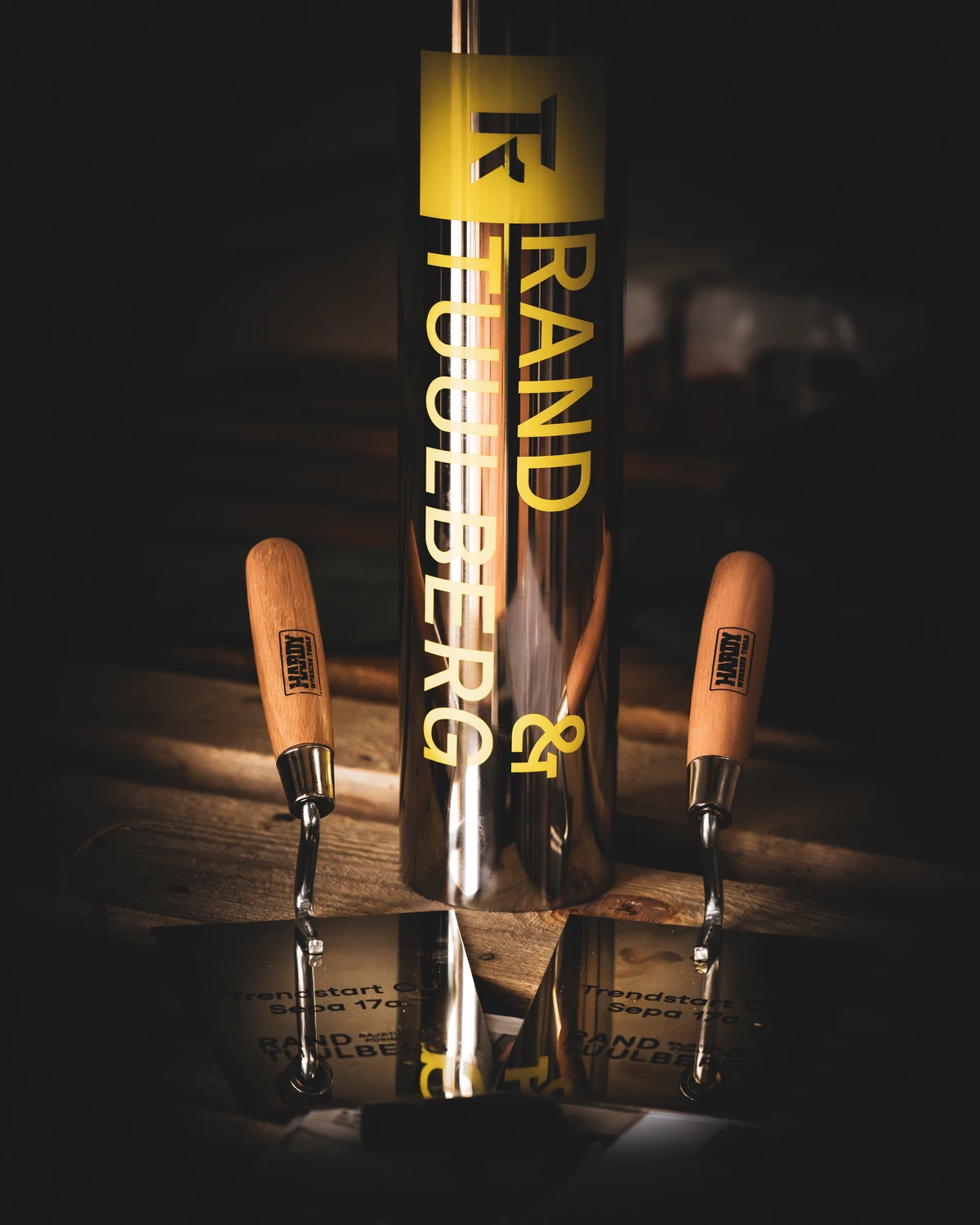 Lähiplaan läikivast nurgakivi kapslist kirjaga "RAND & TUULBERG" ja kahest graveeritud kellust puidust alusel.