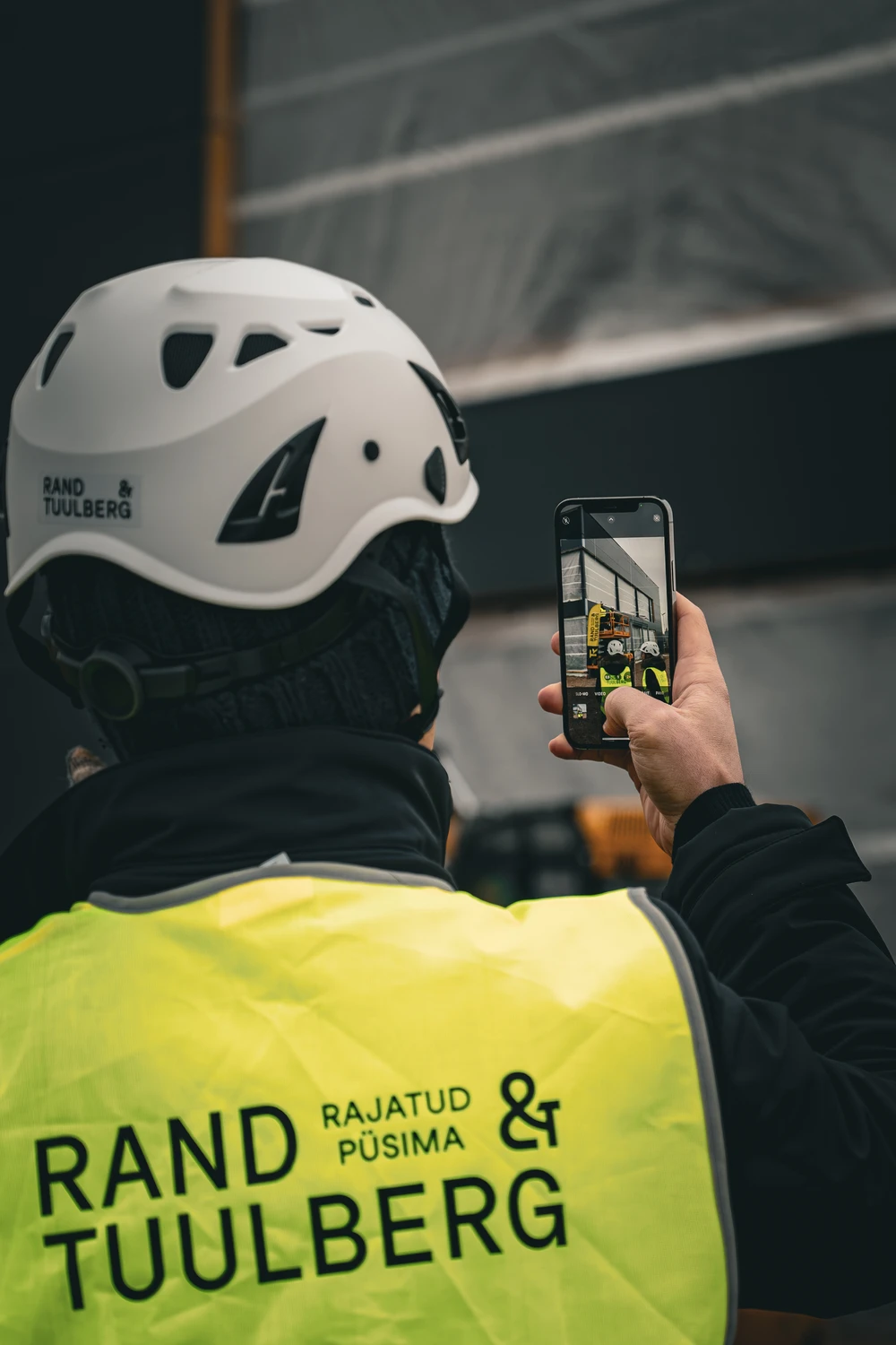 Ehitustööline Rand & Tuulberg logoga kiivriga pildistab nutitelefoniga ehitusplatsil toimuvat.
